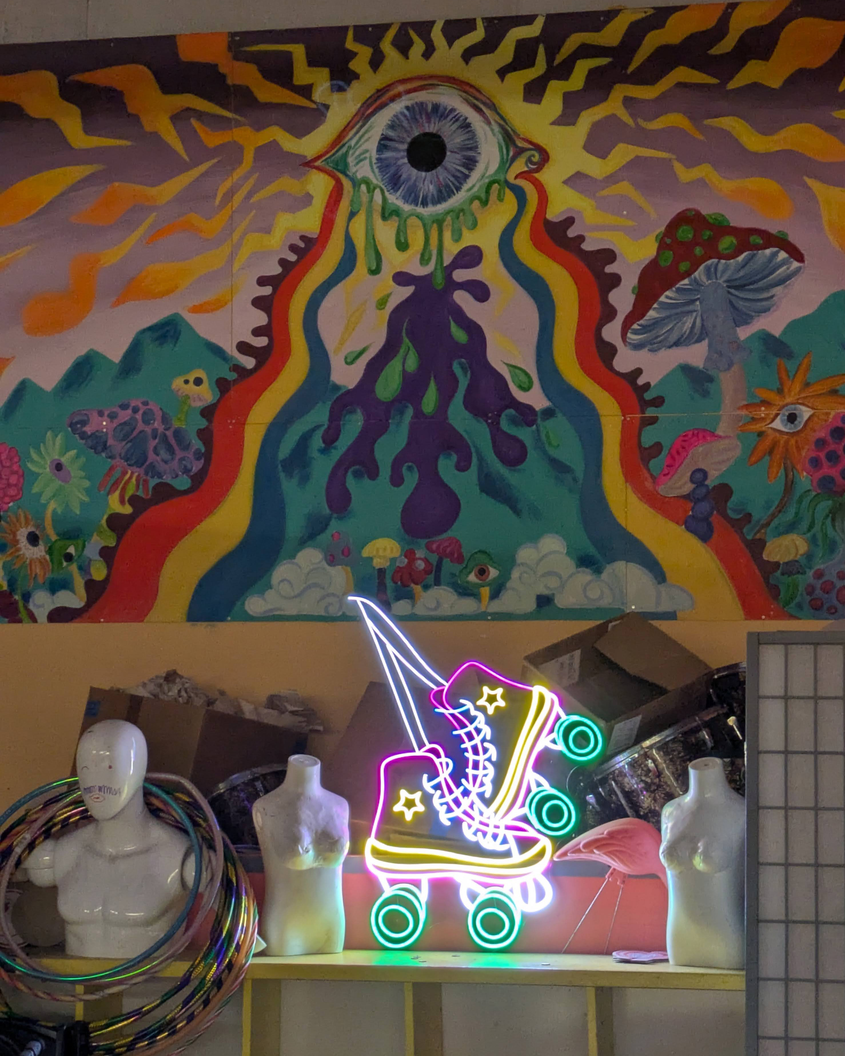 A photo of a neon sign of roller skates on a wall underneath a mural depicting an eye emitting rainbows above a field of mushrooms.